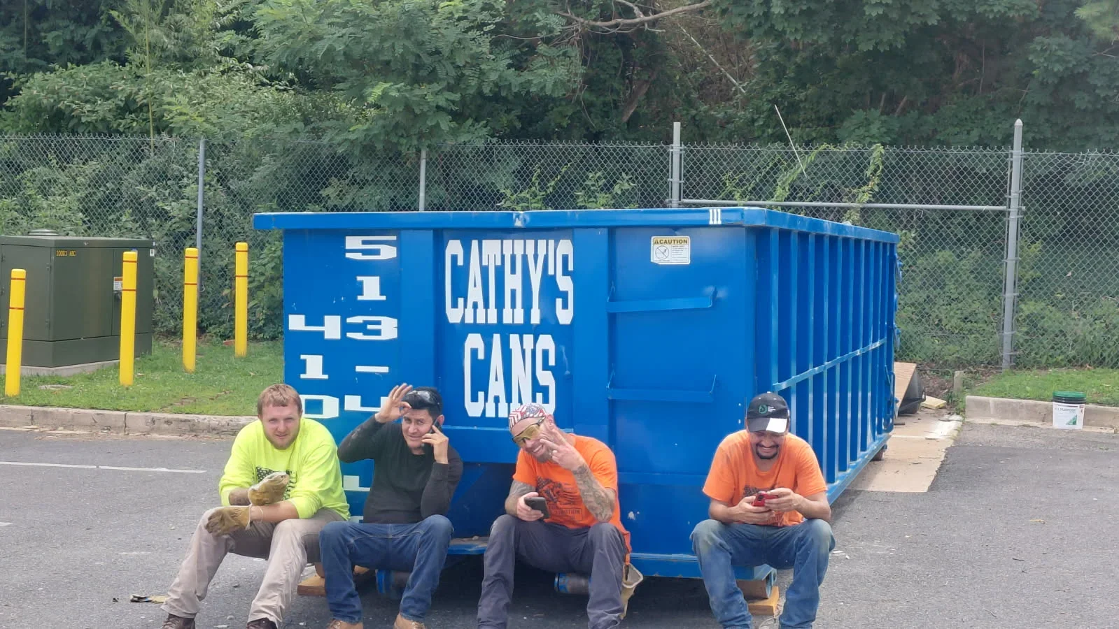 Blue commercial waste container with white branding, steel construction, chain-link fence backdrop