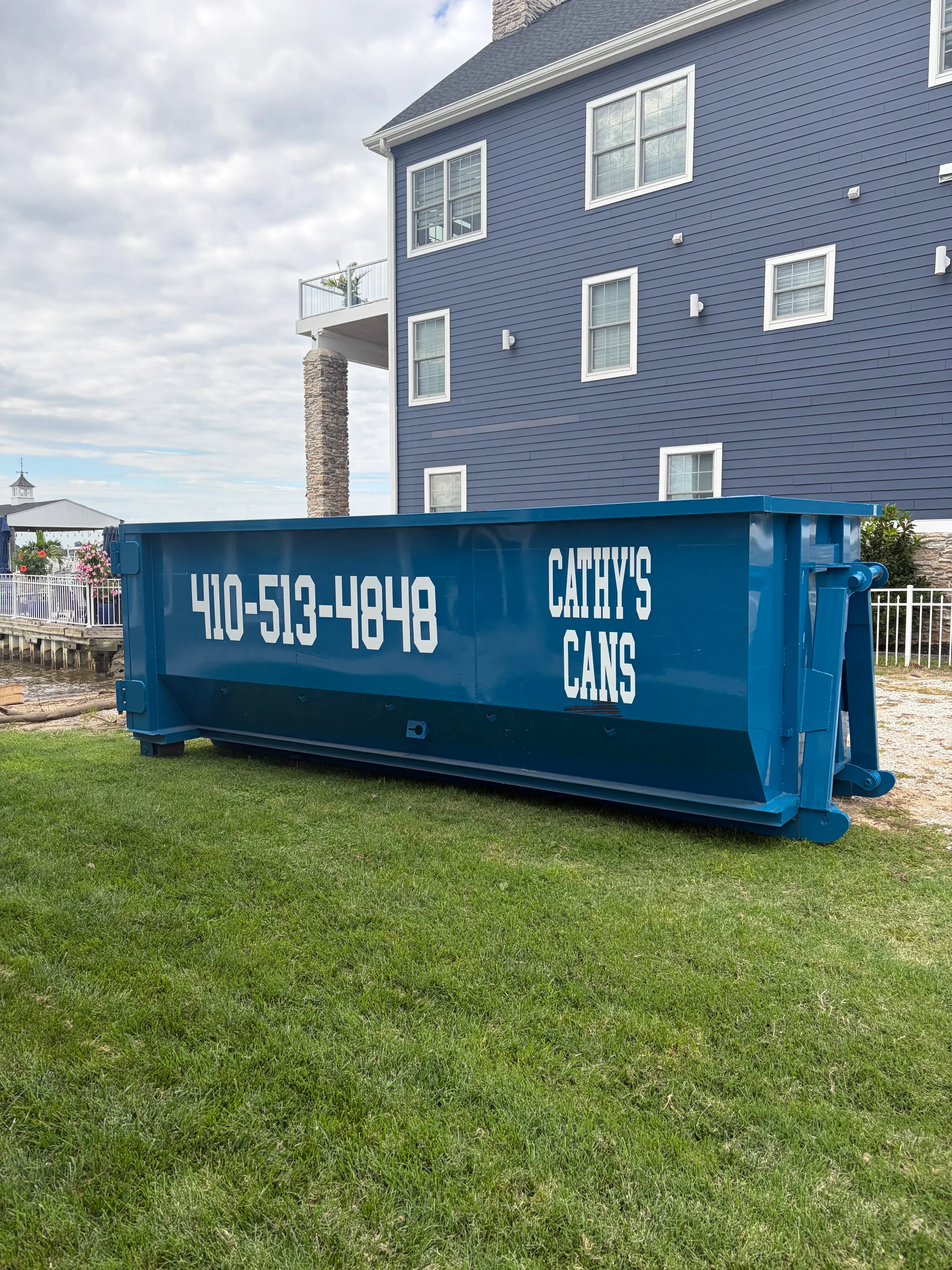 Blue roll-off dumpster with white lettering positioned on residential driveway next to metal-sided home