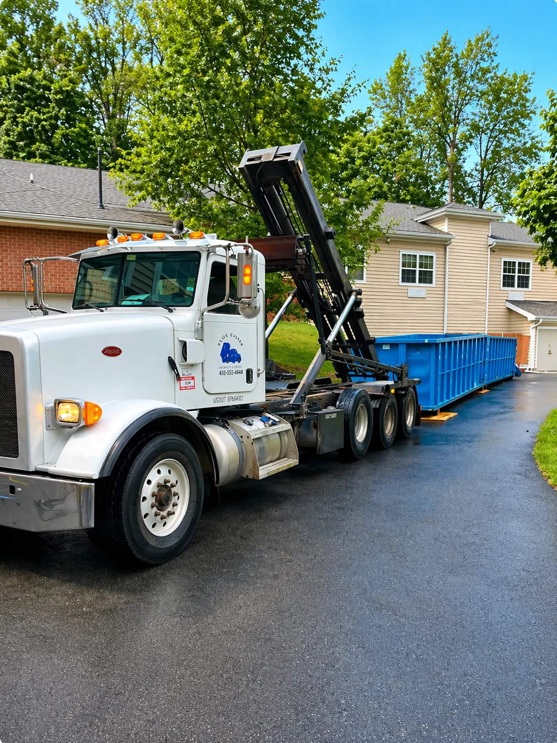 White commercial truck with hydraulic lift lowering blue dumpster container onto residential driveway